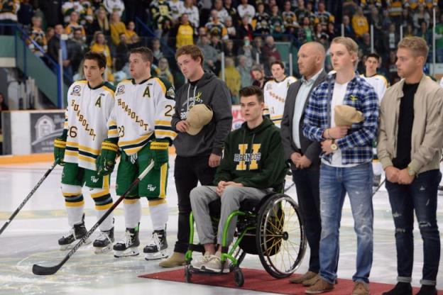 A group of men stand in a hockey rink, wearing shirts and jerseys reading Humboldt Broncos. The one in the centre sits on a wheelchair atop a red carpet.